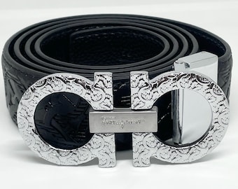 Peut inclure: Une ceinture en cuir noir enroulée en cercle, dotée d'une boucle en métal argenté avec un motif gaufré complexe. La boucle affiche le logo Ferragamo au centre, reliant deux éléments circulaires avec une finition texturée et ornée.