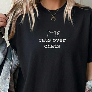 May include: Black t-shirt featuring the white text "cats over chats" and a cat outline. The top has a crew neck. A gold necklace with a crescent moon pendant is worn. The shirt is a casual style.