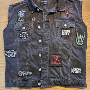 Op de afbeelding: Een zwarte spijkerstof vest met meerdere bandpatches, waaronder "Motorhead England", "Green Day" en "Linkin Park". Het vest heeft een knoopsluiting, twee borstzakken en een kraag. Het vest is mouwloos.
