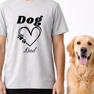 Könnte beinhalten: Hellgraues T-Shirt mit dem schwarzen Schriftzug "Dog Dad" und einem herzförmigen Pfotenabdruck-Design. Ein Golden Retriever-Hund steht neben der Person, die das Shirt trägt.