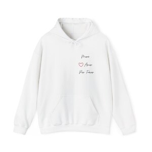 May include: White hoodie with a front pocket and a drawstring hood. The hoodie features the words "More Amor Por Favor" in black script, with a red heart symbol between "Amor" and "Por".