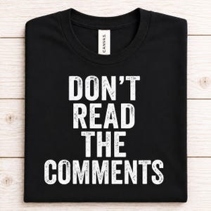 May include: A black short-sleeved t-shirt featuring the white distressed text DON'T READ THE COMMENTS printed in the center. The shirt has a crew neckline and a white brand label attached to the inside of the collar area.