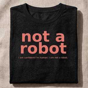 May include: A black crew neck t-shirt featuring the text "not a robot" in large coral-colored letters, with the smaller text "I am confident I'm human. I am not a robot." printed directly underneath in the same coral color.
