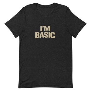 May include: Dark grey t-shirt featuring the text "I'M BASIC" in a tan, slightly distressed font. The shirt has short sleeves and a crew neck. The text is centred on the chest.