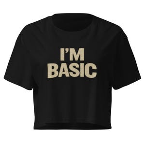 Op de afbeelding: Zwarte crop top met de tekst "I'M BASIC" in een getextureerde, beige lettertype. De top heeft korte mouwen en een ronde hals. Een casual kledingstuk.