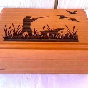 May include: A wooden box with a detailed hunting scene carved on the lid. The design features a hunter aiming a rifle, a dog, flying ducks, and cattails. The box is made of light-colored wood.