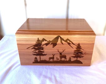 May include: A wooden box with a lid, featuring a laser-etched mountain and forest scene. The design includes trees, deer, and a mountain range. The box is made of light-colored wood with a darker wood lid.