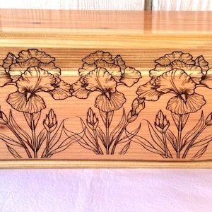 May include: A rectangular wooden box with a curved top. The front of the box features a detailed wood-burned design of three iris flowers and buds. The wood has a light brown colour.