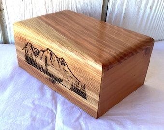 Peut inclure: Une boîte rectangulaire en bois avec un motif de montagne et de forêt gravé sur le côté. La boîte est en bois brun clair avec une finition lisse. La scène de montagne est brun foncé.
