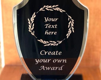 Puede incluir: Un premio de acrílico negro y transparente con forma de escudo. El premio presenta el texto "Your Text here" dentro de una corona de laurel y "Create your own Award" debajo. Se apoya sobre una base negra.