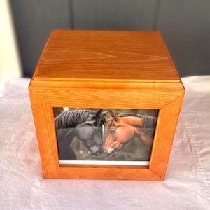 Puede incluir: Una caja de madera cuadrada con un acabado liso de color marrón claro que presenta una fotografía rectangular centrada. La imagen muestra dos caballos, uno negro y otro marrón, posicionados cabeza con cabeza en una interacción suave.
