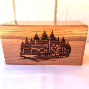 May include: A rectangular wooden box featuring a dark laser-engraved illustration on the front. The design depicts a semi-truck hauling a load of logs, positioned in front of a dense forest of pine trees. The wood grain is visible throughout the surface.