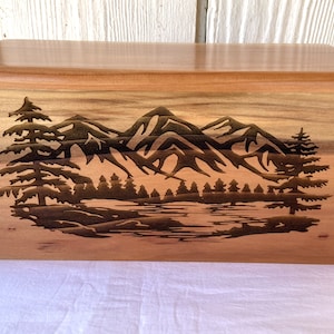 Peut inclure: Une boîte rectangulaire en bois avec une finition au grain brun clair naturel. Le panneau avant présente une illustration de paysage gravée au laser sombre représentant une chaîne de montagnes, un lac calme et des conifères.