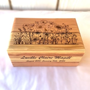 May include: A wooden box with a floral design etched on the lid. The box is light brown with visible wood grain. The name "Lucille Claire Magill" and dates are engraved on the front.