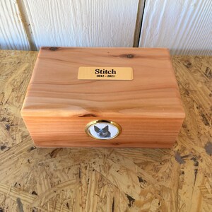 Könnte beinhalten: Eine hölzerne Tierurne mit einer goldenen Namensplakette mit der Aufschrift "Stitch 2012-2021". Auf der Vorderseite der Urne befindet sich ein rundes Foto eines Hundes.