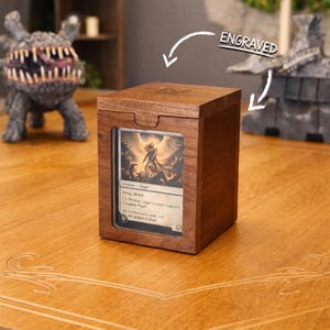 May include: A wooden card storage box with a clear window displaying a card with an angel illustration. The box is engraved with the word "ENGRAVED" and has a sliding lid. The box is sitting on a wooden surface.