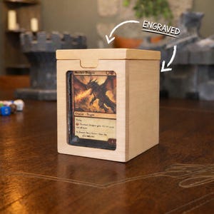 Può includere: Una scatola di legno color chiaro per riporre carte con coperchio scorrevole. La scatola ha una finestra trasparente che mostra una carta con un'illustrazione di drago. La parola "ENGRAVED" è scritta sopra la scatola.