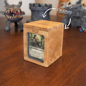 Peut inclure: Une boîte de rangement en bois pour cartes avec une carte visible à l'avant. La boîte est en bois de couleur claire et possède un couvercle à charnière. Le mot "ENGRAVED" est écrit au-dessus de la boîte. La boîte est conçue pour contenir des cartes à collectionner.