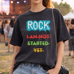 Op de afbeelding: Een zwart t-shirt met de tekst "ROCK I AM NOT STARTED YET" in een kleurrijk ontwerp. Het shirt wordt gecombineerd met een spijkerbroek. Een vrouw draagt het t-shirt op een buitenevenement met een menigte en een reuzenrad op de achtergrond.