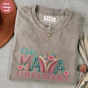 Op de afbeelding: Een grijs t-shirt met zuurwassing met de tekst "This MAMA Loves Books" in teal en roze. Het woord "MAMA" is ontworpen met een gebreid patroon en bloemaccenten. Het shirt heeft een ronde hals en een zilveren ketting.