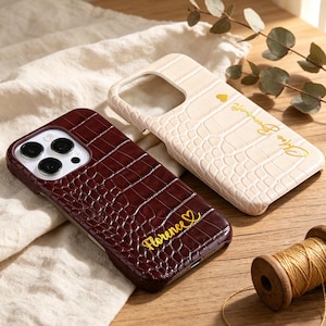 Puede incluir: Dos fundas para smartphone con un patrón de textura de piel de cocodrilo. Una funda es de color burdeos oscuro con el nombre Florence y un símbolo de corazón en letras doradas. La otra es de color crema con un corazón y texto en dorado.