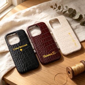 May include: Three phone cases with a crocodile skin texture. One is black with "Chloe Bennett" and a heart, another is burgundy with "Florence" and a heart, and the third is cream-colored with a name and a heart. A spool of gold thread and a gold ring are also visible.