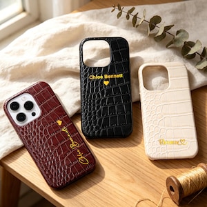 May include: Three phone cases with a crocodile skin texture. One is burgundy, one is black, and one is off-white. Each case has a name and a heart symbol in gold lettering. The cases are on a wooden surface.