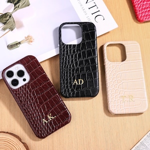 Puede incluir: Tres fundas para teléfono con textura de piel de cocodrilo. Una es burdeos con las iniciales "A.K.", otra es negra con "AD" y la tercera es beige con "T.R.". Las fundas están sobre una superficie de madera clara.