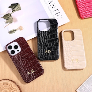 Op de afbeelding: Drie telefoonhoesjes met een krokodillenleertextuur. Eén is bordeauxrood met de initialen "A.K.", één is zwart met "AD" en de derde is beige met "T.R.". De hoesjes liggen op een licht houten oppervlak.
