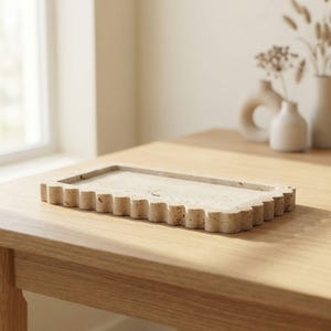 May include: A rectangular, beige stone tray with a scalloped edge sits on a wooden table. The tray has a slightly textured surface and a shallow interior. Decorative vases with dried flowers are in the background.