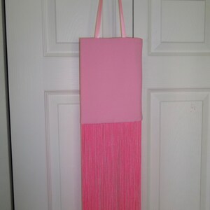 May include: A pink tote bag with a long fringe of pink yarn hanging from the bottom. The bag has pink ribbon straps.