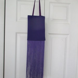 May include: A purple tote bag with a long fringe hanging from the bottom. The bag has a purple strap.
