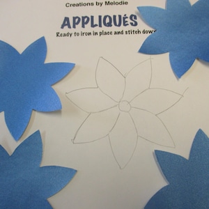 May include: Four blue felt flower appliques and a pencil drawing of a flower on a white background. The text "Appliques" and "Ready to iron in place and stitch down" is printed on the background.