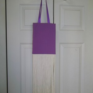 May include: A purple tote bag with a rectangular body and long, thin handles. The bag features a long, white fringe detail at the bottom. The bag is hanging on a white door.