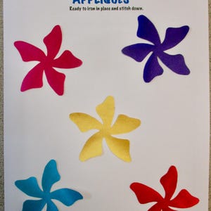 May include: A sheet of paper with five colorful flower appliqués. The appliqués are in shades of pink, purple, yellow, blue, and red. Text at the top reads "APPLIQUÉS" and "Ready to iron in place and stitch down."