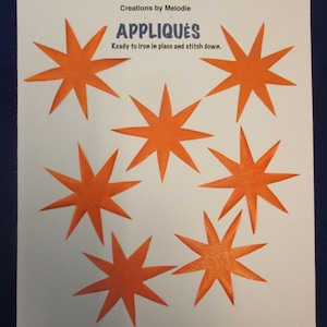May include: Six orange felt star appliques on a white background. The text "Appliques" is printed in blue above the stars. The text "Ready to iron in place and stitch down." is printed in blue below the stars.