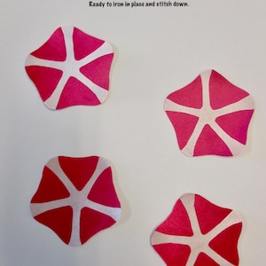 May include: Four appliqués in shades of red and pink, each with a white starburst design. The text "APPLIQUÉS" is in blue, with the phrase "Ready to iron in place and stitch down." The appliqués are ready to be ironed on and stitched.