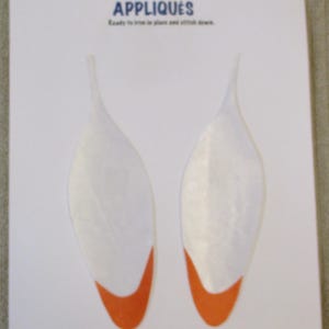 May include: Two white leaf-shaped fabric appliqués with orange tips on a white card. The text at the top reads Creations by Melodie, APPLIQUÉS, and Ready to iron in place and stitch down. The design features two symmetrical, elongated shapes.