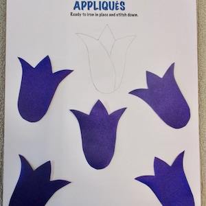May include: A white sheet of paper with the words "Creations by Melodie" and "APPLIQUES" printed in blue. The text "Ready to iron in place and stitch down." is also printed in blue. There are five purple felt flower shapes on the paper. One flower shape is outlined in black.