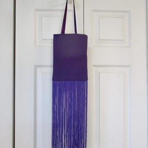May include: A purple tote bag with a long fringe bottom. The bag is hanging from a white hook on a white door.