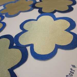 May include: Five gold fabric flower shapes with blue fabric edging. The flowers are arranged on a white surface.