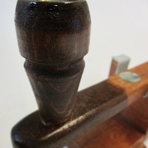 May include: Close-up of a wooden pipe with a dark brown finish. The pipe has a cylindrical bowl and a tapered stem, attached to a wooden base. The wood grain is visible, and the pipe appears to be handcrafted.