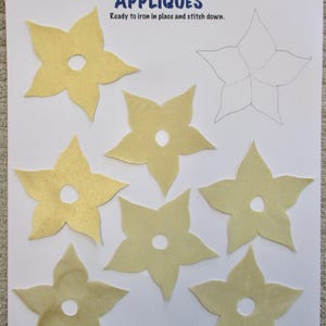May include: A sheet of paper with the text "APPLIQUÉS" and several yellow flower-shaped appliques. The appliques have a hole in the center and are ready to iron on and stitch down. A pencil sketch of a flower is also visible.
