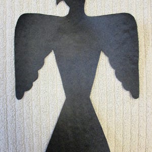 May include: A black felt cutout in the shape of a stylized bird, possibly an eagle or hawk, with outstretched wings. The bird has a detailed head and a flared tail. The cutout is placed on a textured, off-white surface.