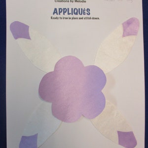 May include: Purple felt flower applique with four white felt feathers. The flower measures 9.25 inches across and the feathers are 5.5 inches long. The text "Creations by Melodie" and "APPLIQUÉS" is printed on the white background. The text "Ready to iron in place and stitch down." is also printed on the background.