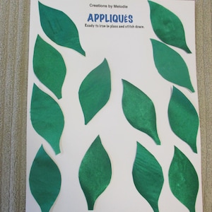 May include: A sheet of white paper with the text "Creations by Melodie APPLIQUES Ready to iron in place and stitch down."  There are ten green felt leaves on the paper.