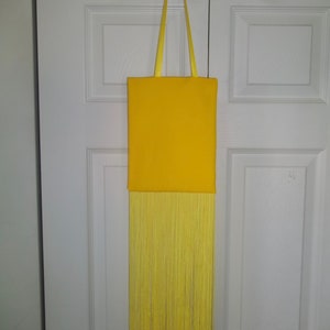 May include: A yellow tote bag with a long fringe hanging from the bottom. The bag has a yellow fabric handle.