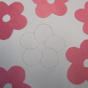 May include: A close-up of a white paper with five pink felt flowers. The flowers have five petals each and a white circle in the center. A sixth flower is drawn in pencil in the center of the image.