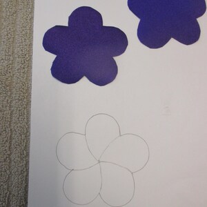 May include: A white sheet of paper with a pencil drawing of a flower with six petals. Three purple felt flowers are shown on the top of the page.
