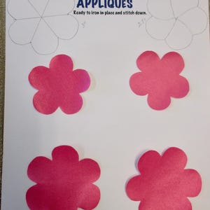 May include: A sheet of paper with the text "APPLIQUÉS" and flower outlines and cutouts. Four pink flower appliqués are displayed, along with two flower outlines. The text "Ready to iron in place and stitch down" is also visible.